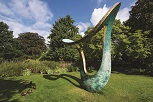 A large, curved copper sculpture illumated by sun. Its shadow curls behind it on a green lawn, with trees in leaf behind.