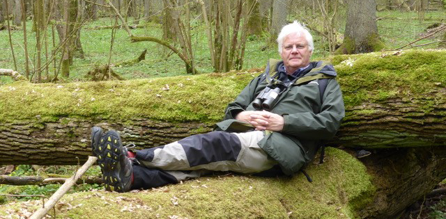 An older man in walking & hiking gear, sitting on a large log which is covered with lichen.