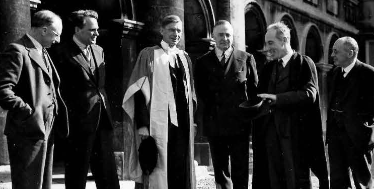 A black & white photograph of men in suits, in the College Cloisters