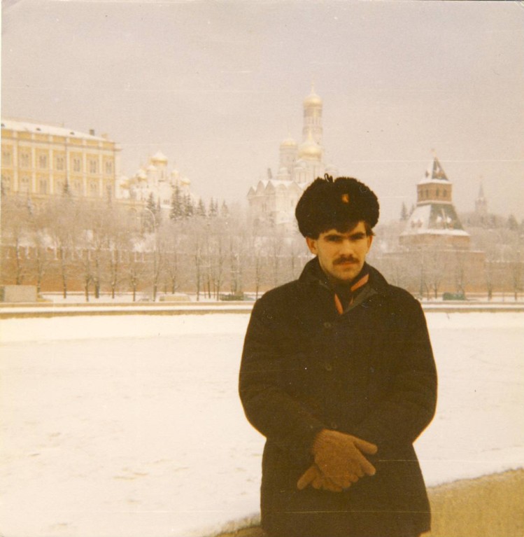 A young man with a dark moustache, standing in front of domed, colourful buildings. There is snow on the ground and he is wearing a large coat, a fur hat and a pink and blue scarf