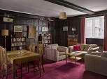 The interior of a dark wood panelled room with a red carpet. Books and paintings on and the walls, a small grey sofa set & a table and chairs on the left hand side. The ceiling is white wood, with a large brown beam across. White-framed clear sash windows with red curtains at the back.