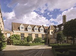 A cobbled square court with paths stretching away from the camera. In the background is a light brick building with white-framed gabled windows in the roof. Small box-hedged flowerbeds around the court with herbs and small trees within.