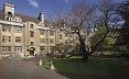 A stone building with large chimneys & doorways, with mullioned windows. Two large trees in leaf in the foreground and a wide pavement area in front.