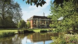 A large pond with trees in leaf surrounding it. In the background a red brick building with stone columns and a central lantern. A green lawn in between and clear blue sky behind.