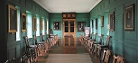 A room with green walls stretching away from the camera. Paintings on all the walls with a glass paned door at the far end, with shields above. A long table and chairs stretch the length of the room.