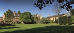 A wide expanse of lawn with trees in leaf. In the background to the left is a large brick building with mullioned windows, the end of a stone building with sash windows and a seventeeth century Chapel with a dark slate roof and cupola. Clear blue sky behind.