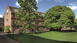 A large brick building with mullioned windows and gables in the brown hanging tiles of the roof. A large lawn in front with trees in leaf along a path.