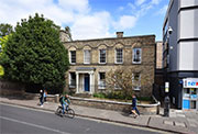 A Georgian lodge building in pale stone-coloured brick with large sash windows along its front and a doorway with a white-columned porch. A large tree in leaf in the foreground.