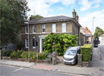 A Georgian house with sash windows and a doorway framed with white columns. A small garden in leaf in front, with a pavement in the foreground.