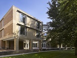 A square, light brick 1960s building with large windows and a terrace in front. A tree in leaf and small lawn in front.