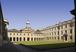 A courtyard with a square lawn, surrounded on three sides by pale stone coloured ranges. They have white framed windows and there is a pediment on the far side. A cupola rises above this, with clear blue sky behind.