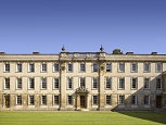 Sunlight illuminating a pale stone-coloured buildings with three levels of white framed sash windows. Two columns either side of three of these windows and a doorway with a crest over it. A neat lawn in front and clear blue sky behind.