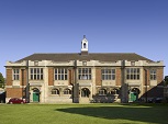 A long brick and stone building with mullioned windows along its first floor. Two green doors at either end and a white lantern in the middle of the grey slate roof. A large expanse of green lawn in front and clear blue sky behind.