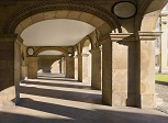 A pale stone coloured cloister illuminated by sun, stretching away from the camera. Memorials on the stones.