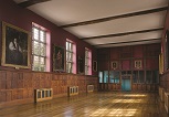 A large wood panelled room stretching away from the camera. Large mullioned windows along the wals, with paintings interspersed between. A medieval screen at the far end, with shields placed above. The floor is wooden and it is illumated by sun from the windows.