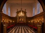 Chapel interior stretching away from the camera. The pavement is black and white and the room is half wood panelled. The rest of the room is white, with an altarpiece and rail at the far end. There is a large glass chandelier in the middle, and the image is illuminated with candles.