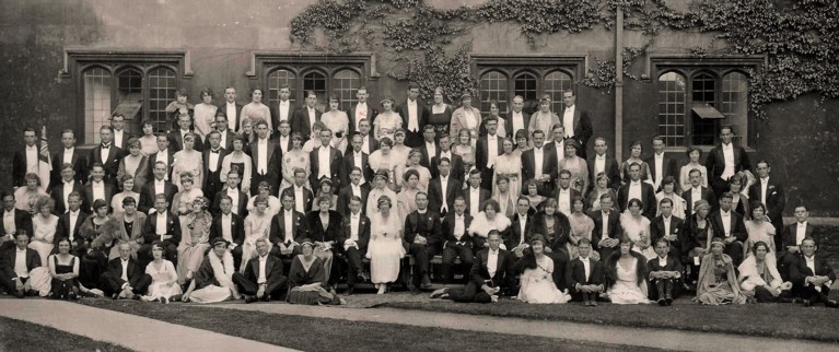 A black and white photograph of formally dressed men and women, sitting in front of a wall and windows