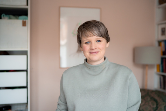 A young woman with short greying hair, standing in a comfortable office. She has blue eyes and is wearing a pale green–grey polo neck jumper.