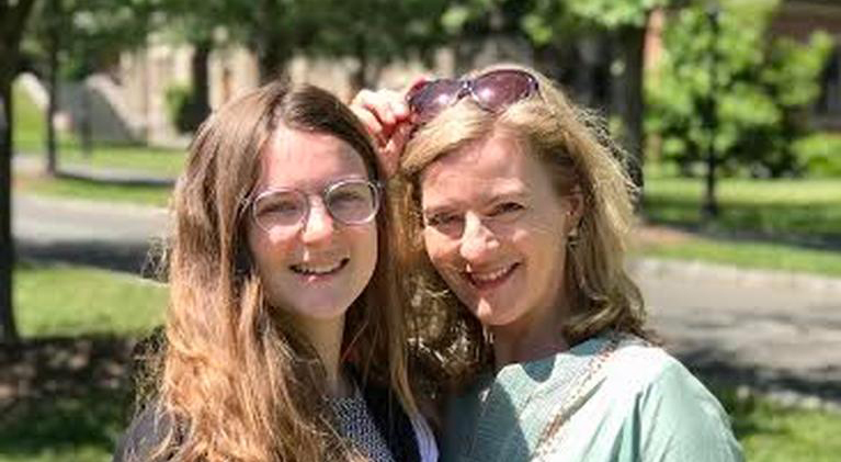 Two women standing close together: left–hand woman is in her early 20s, has glasses and has long brown hair, the right–hand woman is in her 40s and has blond hair. They are standing in the sun and smiling.