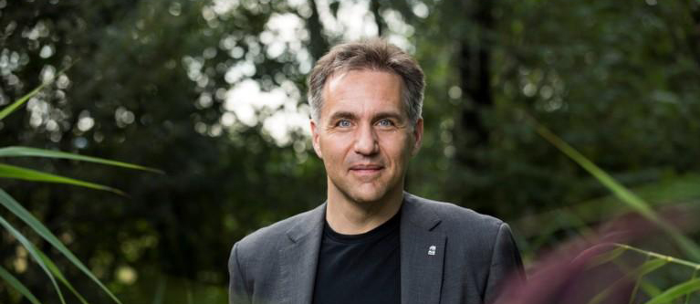 A man in his 40s standing in tropical foliage. He has greying hair, blue eyes and tanned skin, and he is wearing a black t–shirt & grey jacket with a WWF pin of a panda on his lapel.