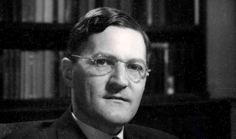 A black & white photo of a middle-aged man wearing glasses, with a bookcase behind.