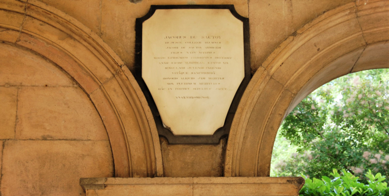 A plaque on the College's cloister, with arches & leaves in the background