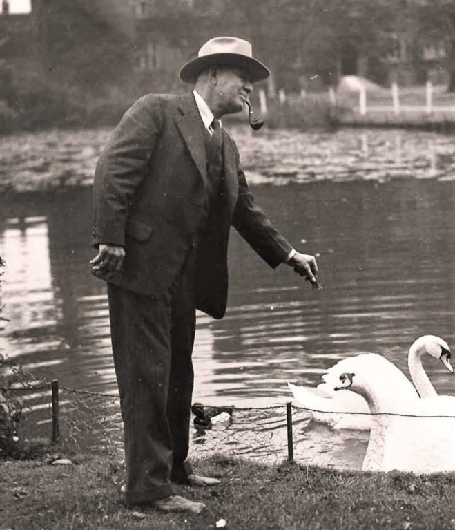 A black and white photograph of a middle-aged man in a suit and hat, with a pipe in his mouth, standing by a pond