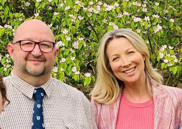 A middle-aged bald man in glasses standing next to a blond woman in a pink shirt and jacket, standing in front of a tree with white flowers on