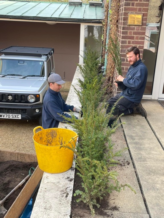 Two men in blue jackets planting a hedge next to a brick house