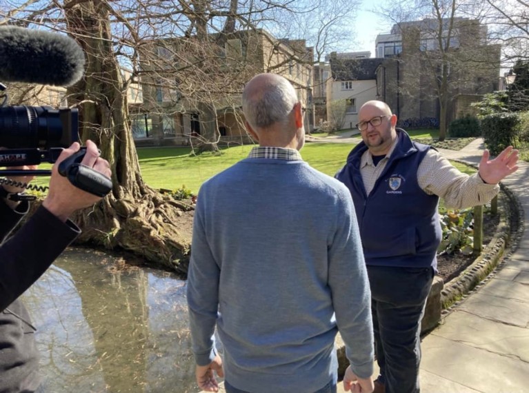 A middle-aged man interviewing another man, who is wearing a blue Emmanuel College-branded jacket, with gardens and a small pond in the background