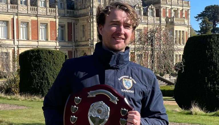 A young man in an Emmanuel College-branded jacket standing in front of a large red-brick house and lawns, holding a shield