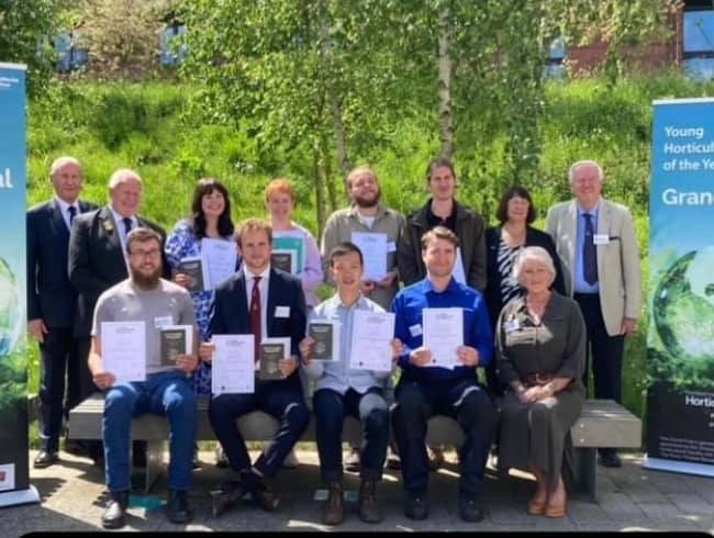 A group of men and women seated and standing, holding certificates, with trees behind