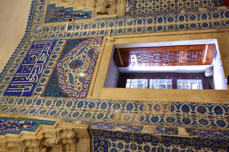 A wall of blue, white and red Iznik tiles, with flowers, leaves and Arabic letters. The rest of the walls are carved marble