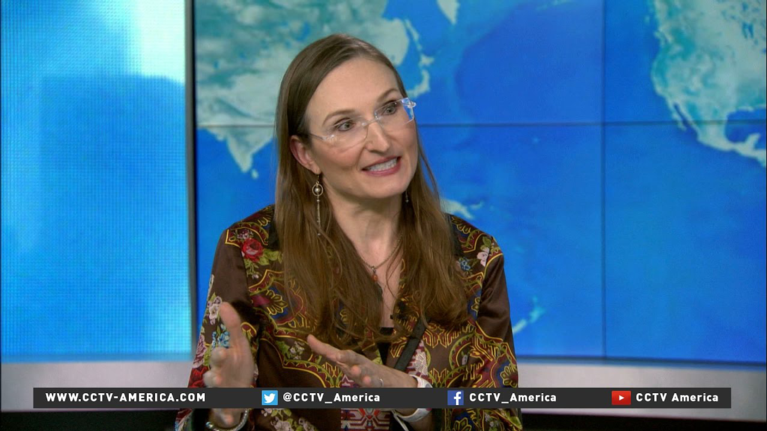 A screengrab of a middle–aged woman with long brown hair speaking to a camera, with a map of the world on a screen behind her.