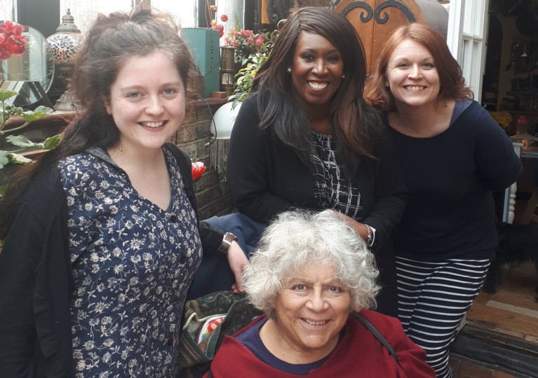 Three young women grouped around a seated middle-aged woman with white hair. The woman on the left is white, with long brown hair and a heart-shaped face. The woman in the centre is a person of colour, is dressed in black, with long black hair. The woman to the right-hand side is dressed in blue, with red hair. They are all smiling at the camera.