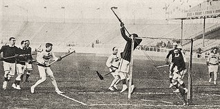 A black and white newspaper clipping of a group of men playing lacrosse in a small stadium