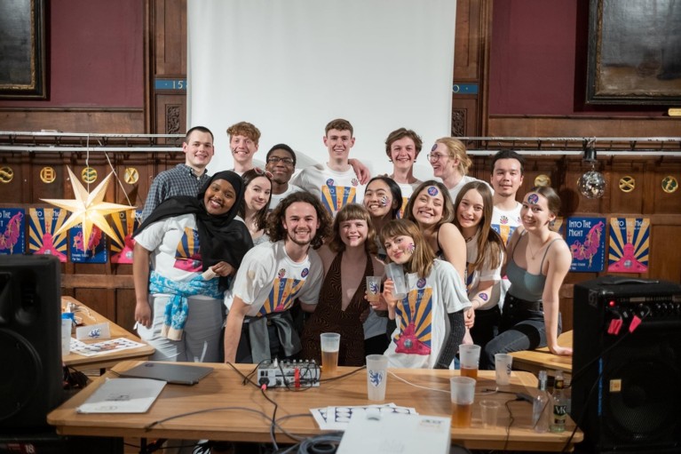 A group of students posing in front of a line of May Ball posters