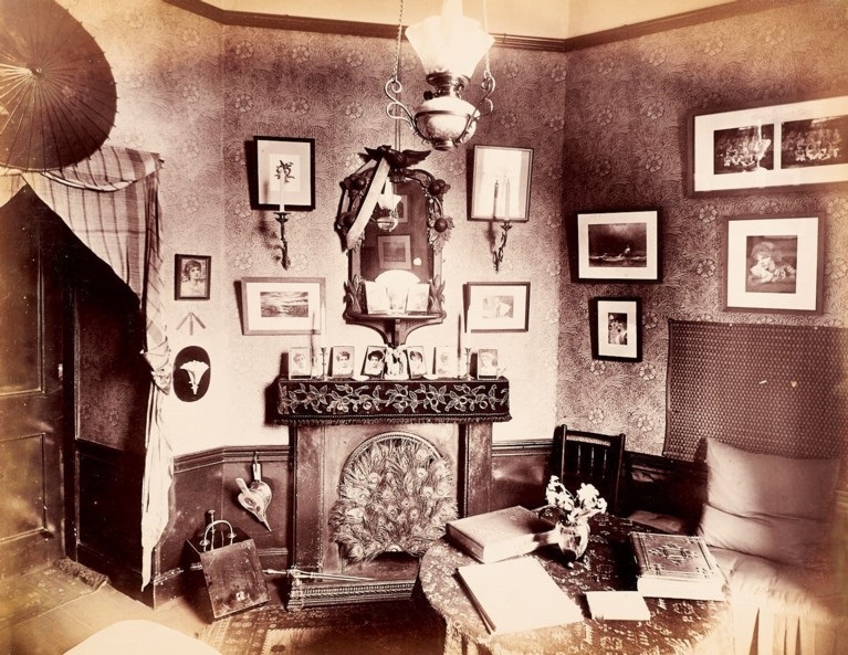 A black and white photograph of the inside of a college room with ornate fireplace, table & chairs and pictures on the walls