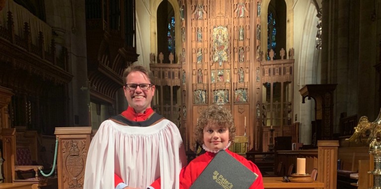A young boy in a red cassock, holding a music folder, next to a middle-aged man with glasses, also in a red cassock, standing in a church