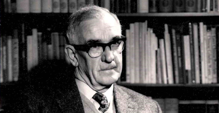 An elderly man sitting in front of a bookcase, wearing a suit and thick-rimmed black glasses