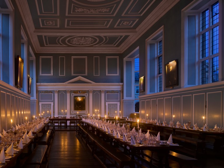 A candlelit blue and white pannelled hall with long brown benches and tables, laid for dinner 