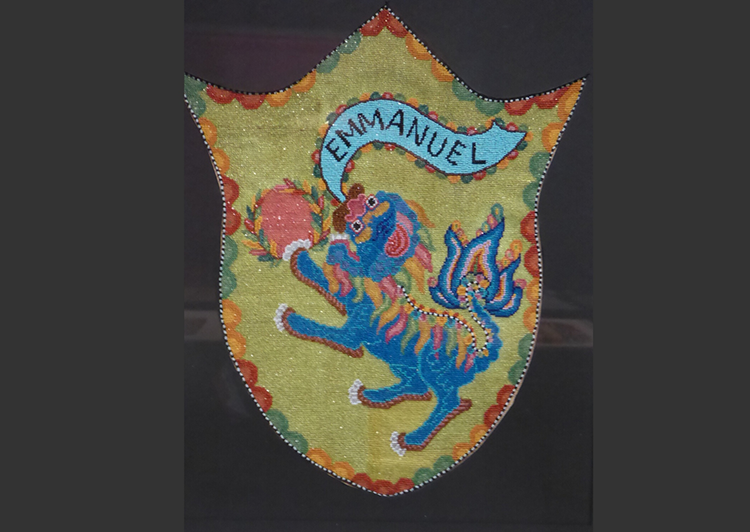 A traditional Peranakan–style bead panel: a blue Chinese Foo dog, with a based on the Emmanuel lion emblem, becoming a Chinese Foo dog, with a banner flowing from its mouth reading 'Emmanuel', and a flaming pearl. 