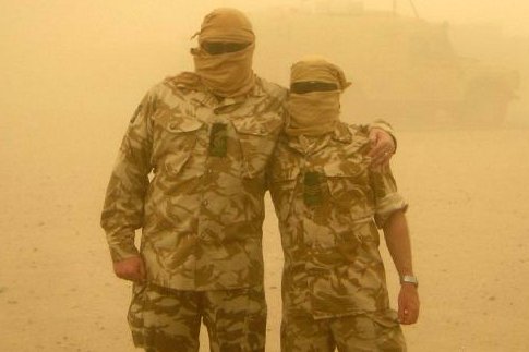 Two men in army fatigues, face coverings, and glasses, in a sandstorm.